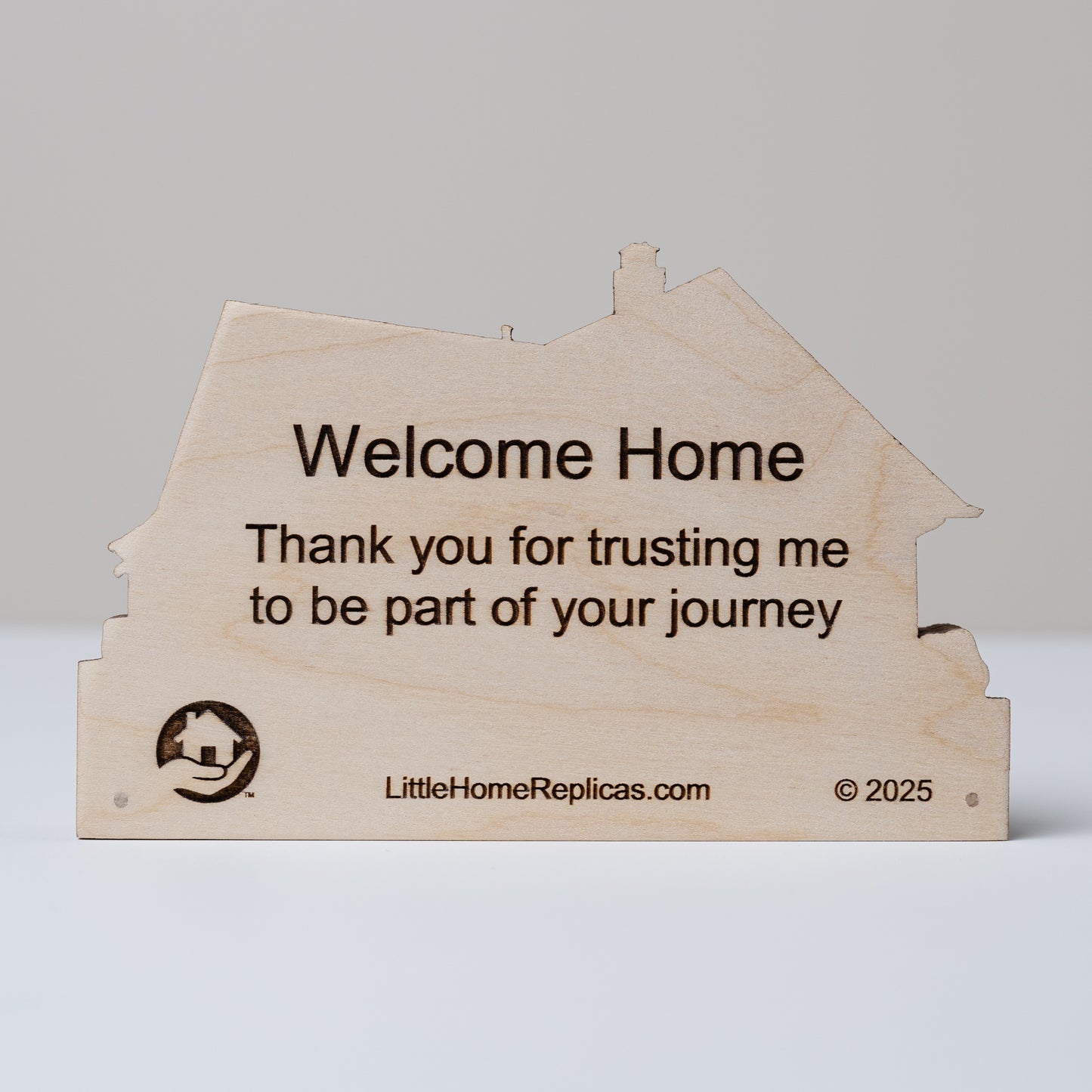 Wooden house-shaped replica with 'Welcome Home' and 'Thank you for trusting me to be a part of your journey' message on a gray background