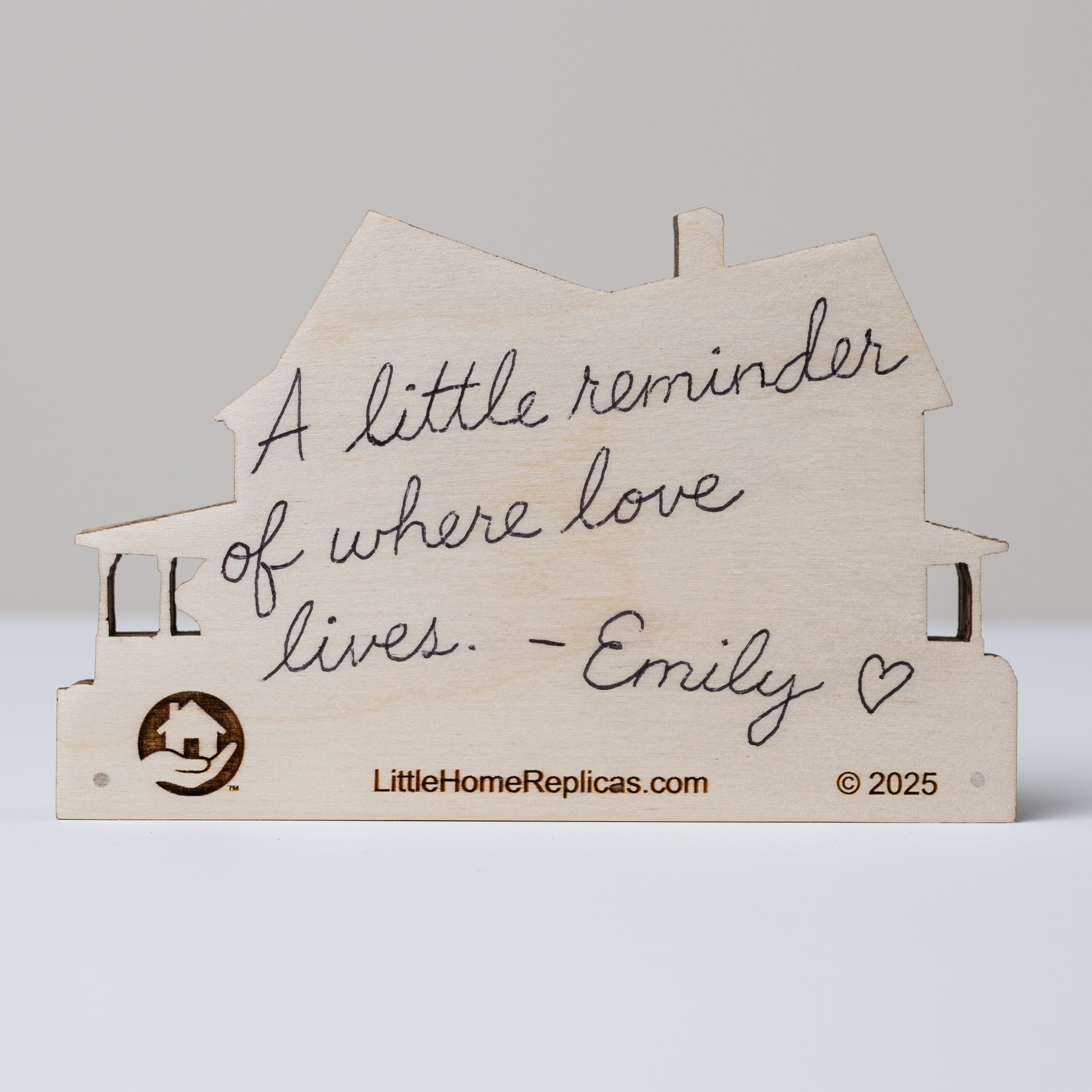 Wooden house-shaped replica with handwritten text 'a little reminder of where love lives. -Emily' on a gray background.